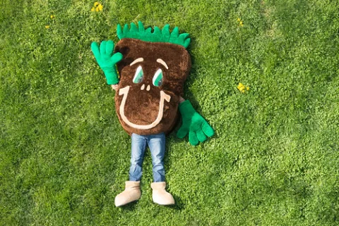 Sammy Soil mascot lays on a field of grass and waves up at the camera.