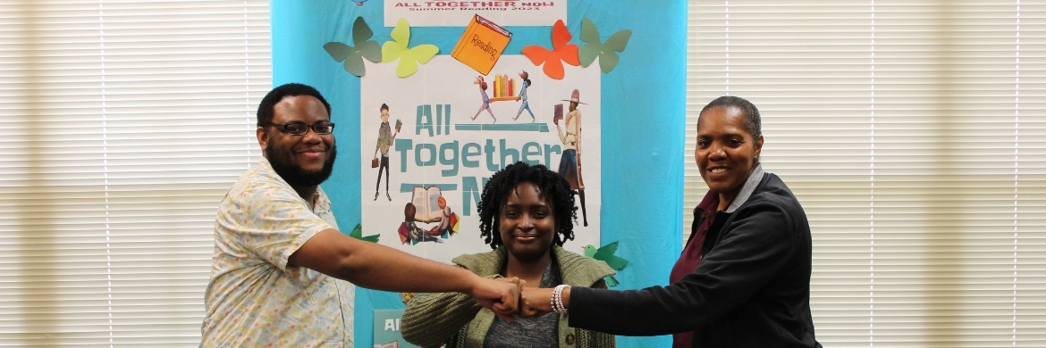 Three adults smiling and fist bumping inside the library during a reading event